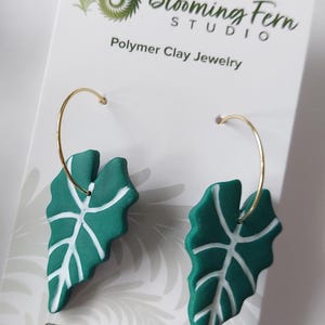 May include: A pair of hoop earrings featuring dark green polymer clay leaf pendants with white vein details. The earrings are attached to a white display card printed with the text Blooming Fern Studio Polymer Clay Jewelry and social media info.
