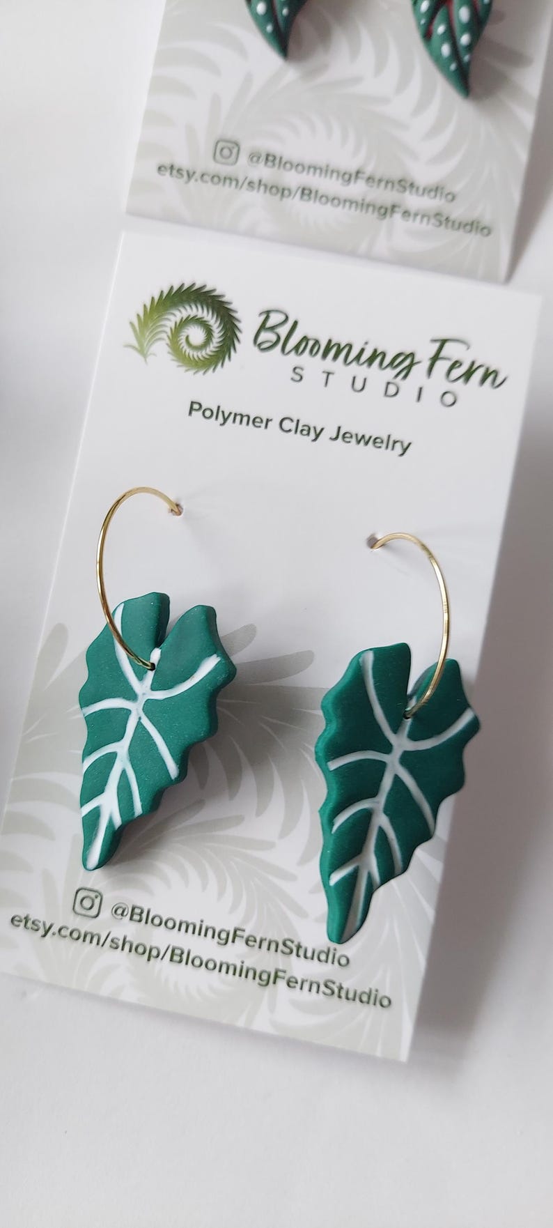 May include: A pair of hoop earrings featuring dark green polymer clay leaf pendants with white vein details. The earrings are attached to a white display card printed with the text Blooming Fern Studio Polymer Clay Jewelry and social media info.