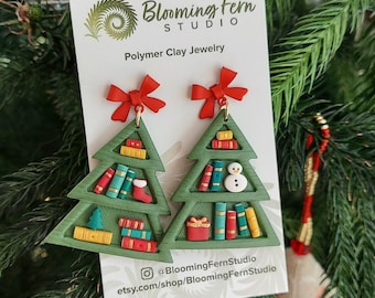 May include: Green polymer clay earrings shaped like Christmas trees, adorned with miniature books, a snowman, and a gift. Each tree has a red bow at the top. The earrings are displayed on a white card with the Blooming Fern Studio logo.