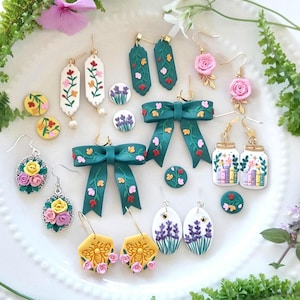 May include: A collection of handmade earrings in various designs, including floral, bow, and bee motifs. The earrings feature vibrant colors like green, pink, yellow, and purple. Some earrings have a gold-colored metal finish. The earrings are displayed on a white plate.
