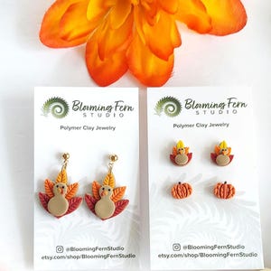 May include: Polymer clay jewelry featuring turkey and pumpkin designs. The earrings and studs are displayed on white cards with the Blooming Fern Studio logo and Etsy shop URL. An orange flower and decorative beads are also in the image.