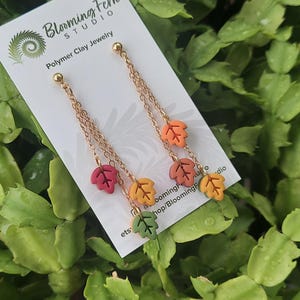 May include: Gold-colored chain earrings with colorful polymer clay leaf charms. The leaves are in shades of red, yellow, orange, and green. The earrings are displayed on a white card with the text "Blooming Fern Studio Polymer Clay Jewelry".