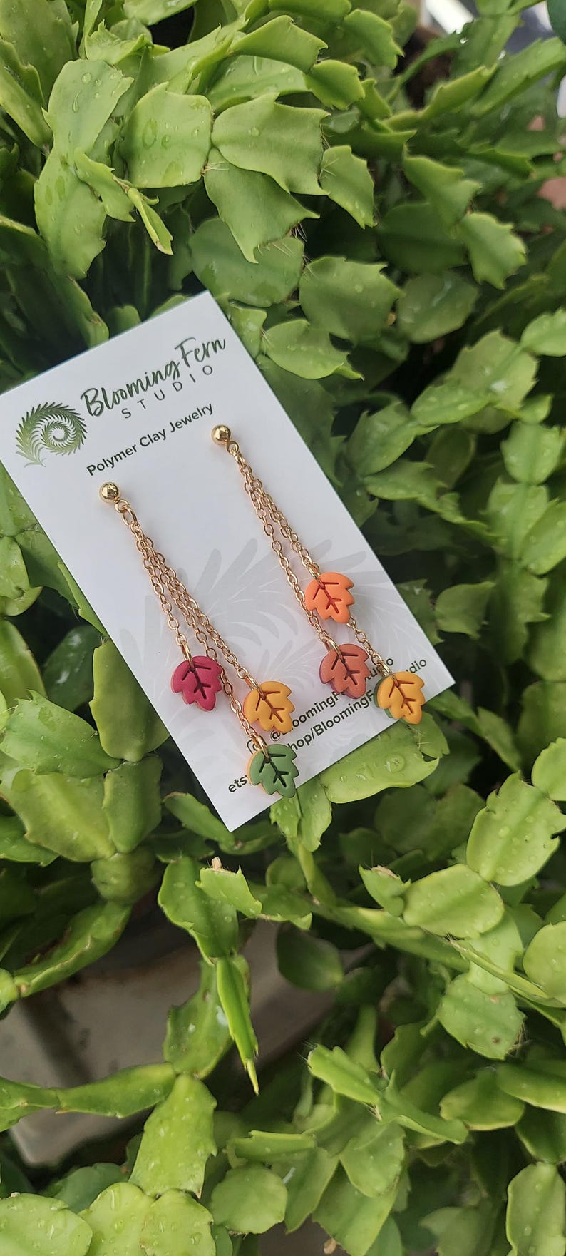 May include: Gold-colored chain earrings with colorful polymer clay leaf charms. The leaves are in shades of red, yellow, orange, and green. The earrings are displayed on a white card with the text "Blooming Fern Studio Polymer Clay Jewelry".