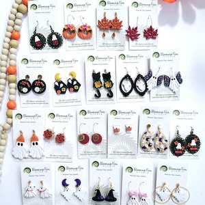 May include: A collection of polymer clay earrings with various Halloween and autumn themes. Designs include ghosts, pumpkins, cats, and fall leaves. The earrings are displayed on white cards with the Blooming Fern Studio logo.