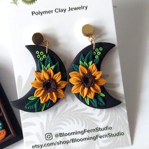 May include: A pair of black crescent moon-shaped polymer clay earrings featuring yellow sunflowers with brown centers and green leaves. The earrings are attached to a white display card with the text Blooming Fern Studio Polymer Clay Jewelry.