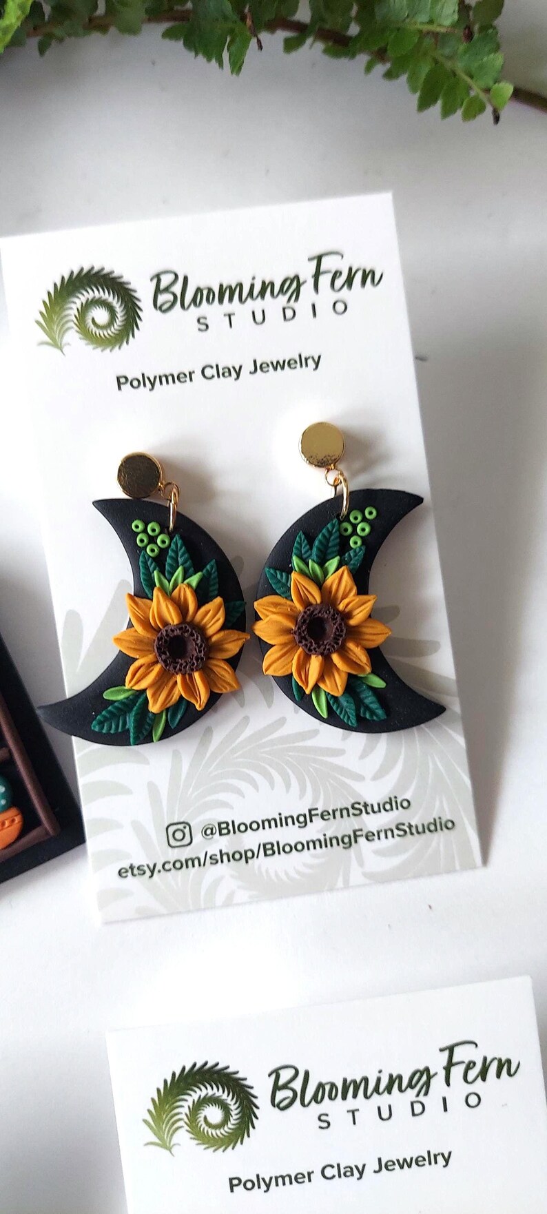 May include: A pair of black crescent moon-shaped polymer clay earrings featuring yellow sunflowers with brown centers and green leaves. The earrings are attached to a white display card with the text Blooming Fern Studio Polymer Clay Jewelry.