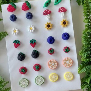 May include: A collection of small stud earrings arranged on a white card, featuring various miniature food and nature designs including raspberries, blueberries, strawberries, blackberries, peas, mushrooms, sunflowers, daisies, and citrus fruit slices.
