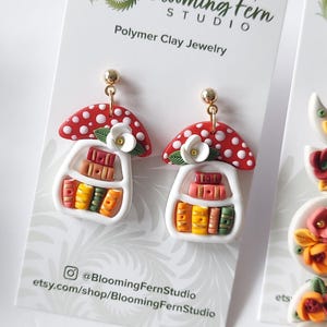 May include: A pair of polymer clay earrings featuring a red mushroom cap with white spots atop a white bookshelf filled with miniature colorful books. A small white flower with green leaves sits on the shelf. The earrings have gold-tone posts.