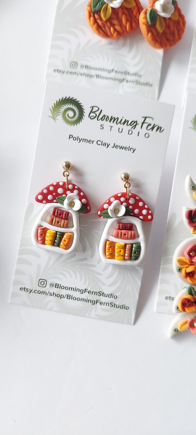 May include: A pair of polymer clay earrings featuring a red mushroom cap with white spots atop a white bookshelf filled with miniature colorful books. A small white flower with green leaves sits on the shelf. The earrings have gold-tone posts.