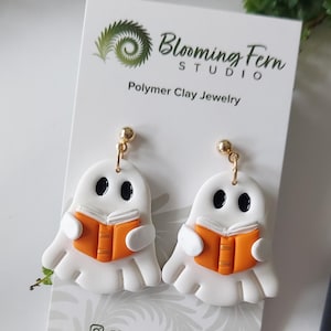May include: A pair of white ghost-shaped polymer clay earrings featuring black oval eyes and holding an orange book with white pages. The earrings are attached to a white display card with the text Blooming Fern Studio Polymer Clay Jewelry.