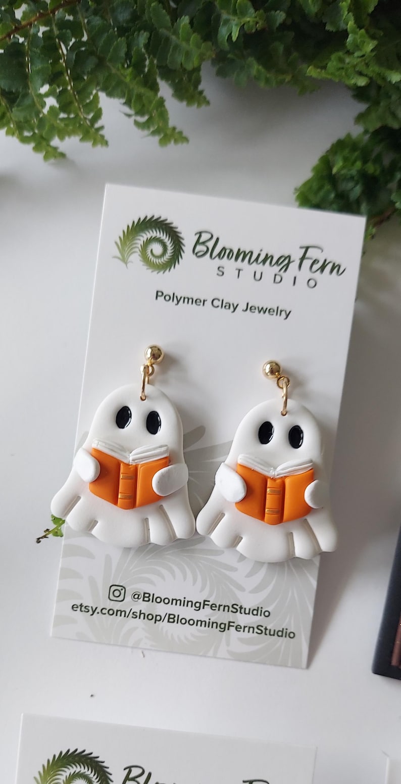 May include: A pair of white ghost-shaped polymer clay earrings featuring black oval eyes and holding an orange book with white pages. The earrings are attached to a white display card with the text Blooming Fern Studio Polymer Clay Jewelry.