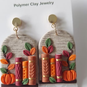 May include: A pair of arch-shaped polymer clay earrings featuring a textured beige background with three miniature books in shades of red, orange, and brown, accented with gold details, green and orange leaves, and a small orange pumpkin at the base.