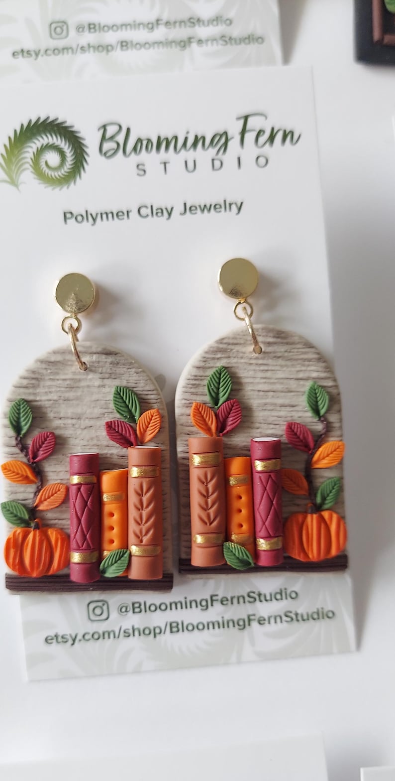 May include: A pair of arch-shaped polymer clay earrings featuring a textured beige background with three miniature books in shades of red, orange, and brown, accented with gold details, green and orange leaves, and a small orange pumpkin at the base.
