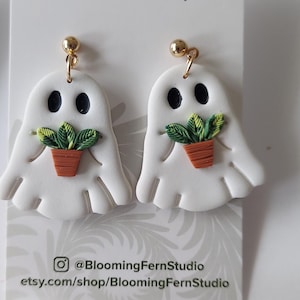 May include: A pair of white ghost-shaped polymer clay earrings featuring black eyes and a small green potted plant held in the center. The earrings are attached to gold-toned metal posts and are displayed on a white Blooming Fern Studio card.