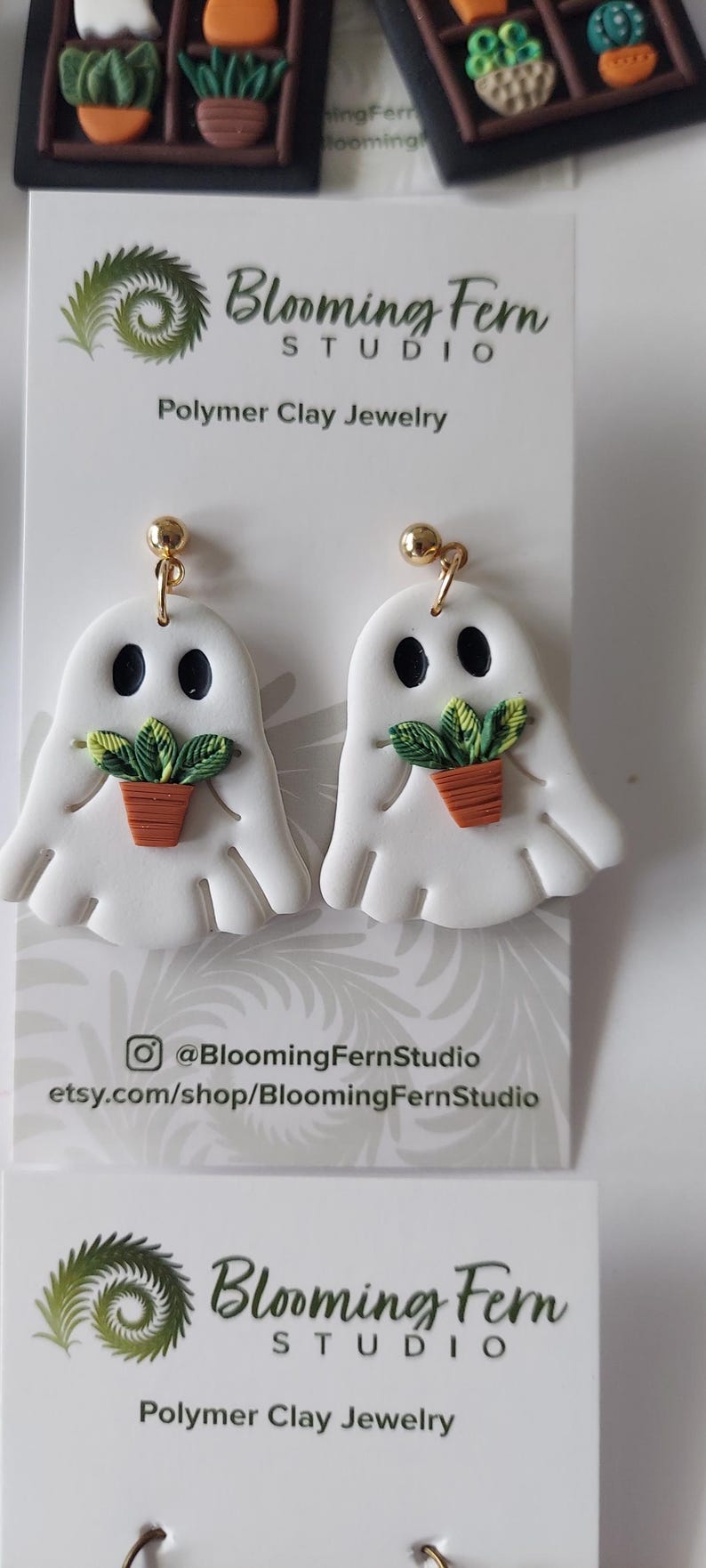 May include: A pair of white ghost-shaped polymer clay earrings featuring black eyes and a small green potted plant held in the center. The earrings are attached to gold-toned metal posts and are displayed on a white Blooming Fern Studio card.