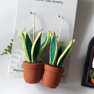 May include: A pair of dangle earrings featuring miniature potted snake plants made of polymer clay. The plants have green leaves with yellow edges, set in terracotta-colored pots. The earrings are attached to a white display card with the text Blooming Fern Studio Polymer Clay Jewelry.