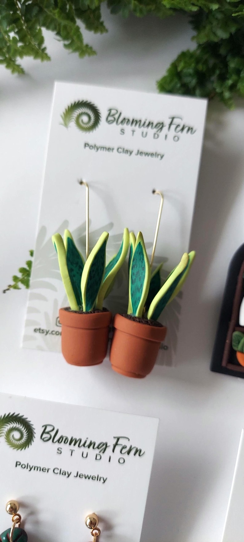 May include: A pair of dangle earrings featuring miniature potted snake plants made of polymer clay. The plants have green leaves with yellow edges, set in terracotta-colored pots. The earrings are attached to a white display card with the text Blooming Fern Studio Polymer Clay Jewelry.