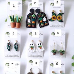 May include: A collection of nine pairs of polymer clay earrings displayed on white cards, featuring designs such as potted snake plants, arched windows with ghosts and plants, sunflowers on crescent moons, leaf shapes, ghosts holding plants, and snakes.
