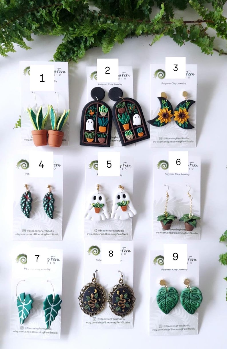 May include: A collection of nine pairs of polymer clay earrings displayed on white cards, featuring designs such as potted snake plants, arched windows with ghosts and plants, sunflowers on crescent moons, leaf shapes, ghosts holding plants, and snakes.