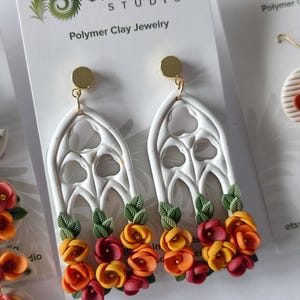 May include: Dangle earrings featuring white polymer clay gothic arch frames adorned with small sculpted orange, yellow, and red flowers and green leaves. The earrings have gold-toned circular studs and are displayed on a white Blooming Fern Studio card.