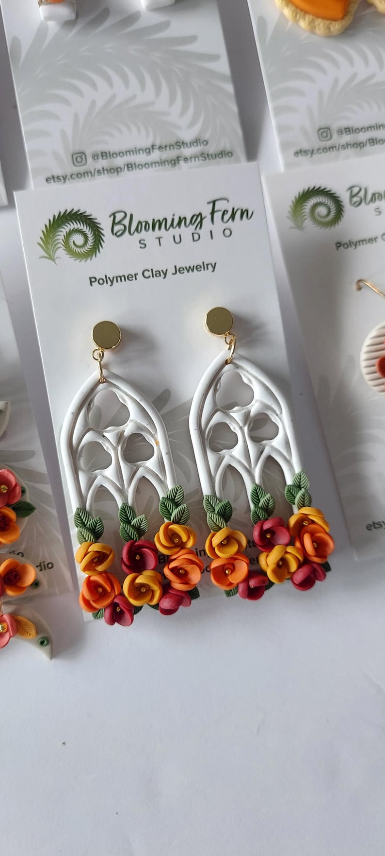 May include: Dangle earrings featuring white polymer clay gothic arch frames adorned with small sculpted orange, yellow, and red flowers and green leaves. The earrings have gold-toned circular studs and are displayed on a white Blooming Fern Studio card.