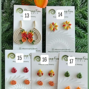 May include: A collection of polymer clay earrings in various fall-themed designs. The earrings include leaf, pumpkin, and flower shapes in shades of red, orange, yellow, and green. Some earrings have a floral design with red and orange flowers. The Blooming Fern Studio logo is visible.