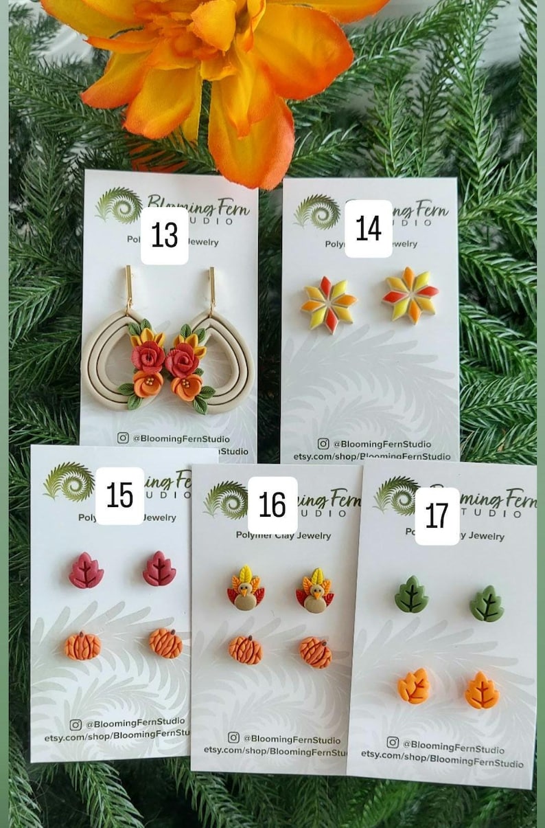 May include: A collection of polymer clay earrings in various fall-themed designs. The earrings include leaf, pumpkin, and flower shapes in shades of red, orange, yellow, and green. Some earrings have a floral design with red and orange flowers. The Blooming Fern Studio logo is visible.