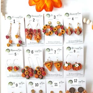May include: A collection of polymer clay earrings with fall and autumn themes. Designs include pumpkins, leaves, sunflowers, and turkey shapes. The earrings are in various colors, including orange, red, and yellow. The brand name "Blooming Fern Studio" is visible on the tags.