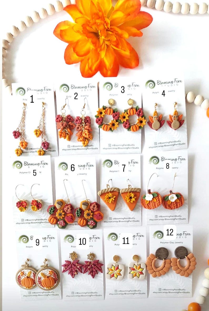 May include: A collection of polymer clay earrings with fall and autumn themes. Designs include pumpkins, leaves, sunflowers, and turkey shapes. The earrings are in various colors, including orange, red, and yellow. The brand name "Blooming Fern Studio" is visible on the tags.