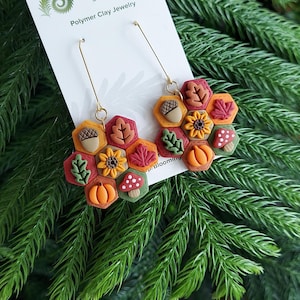 May include: Polymer clay earrings featuring a fall theme. The earrings have a honeycomb design with autumn leaves, acorns, pumpkins, and mushrooms. The earrings are on a white card with the text "Blooming Fern Studio Polymer Clay Jewelry".