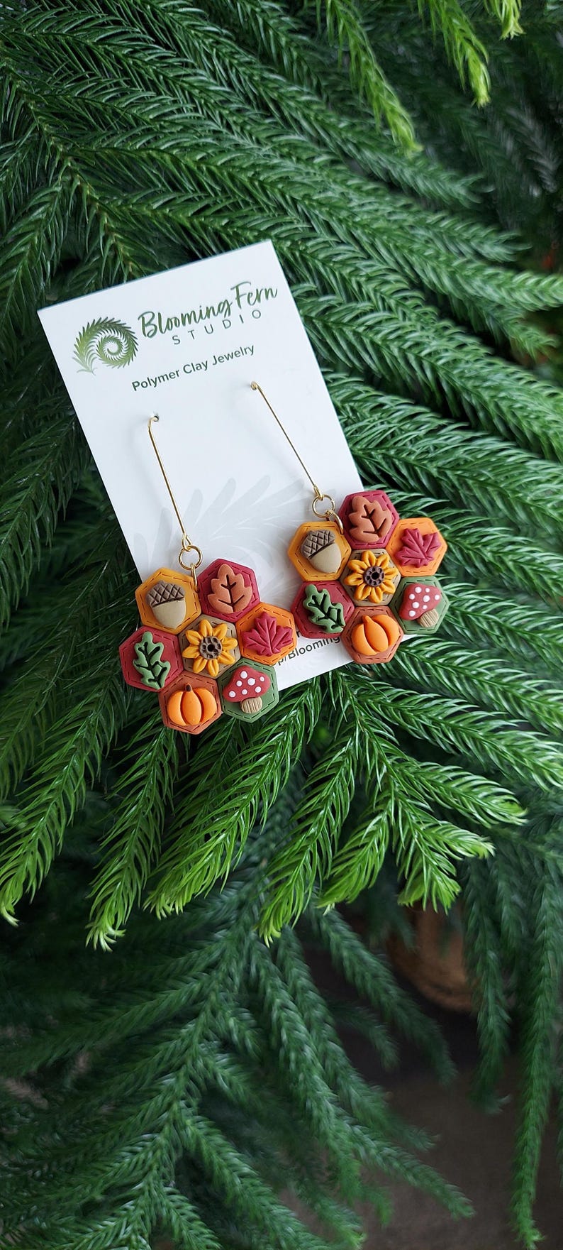 May include: Polymer clay earrings featuring a fall theme. The earrings have a honeycomb design with autumn leaves, acorns, pumpkins, and mushrooms. The earrings are on a white card with the text "Blooming Fern Studio Polymer Clay Jewelry".