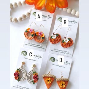 May include: A selection of polymer clay earrings in various designs. The earrings include orange pumpkin hoops, pumpkin-shaped earrings with white flowers, teardrop earrings with floral accents, and pie-shaped earrings with sunflowers. The earrings are displayed on white cards.