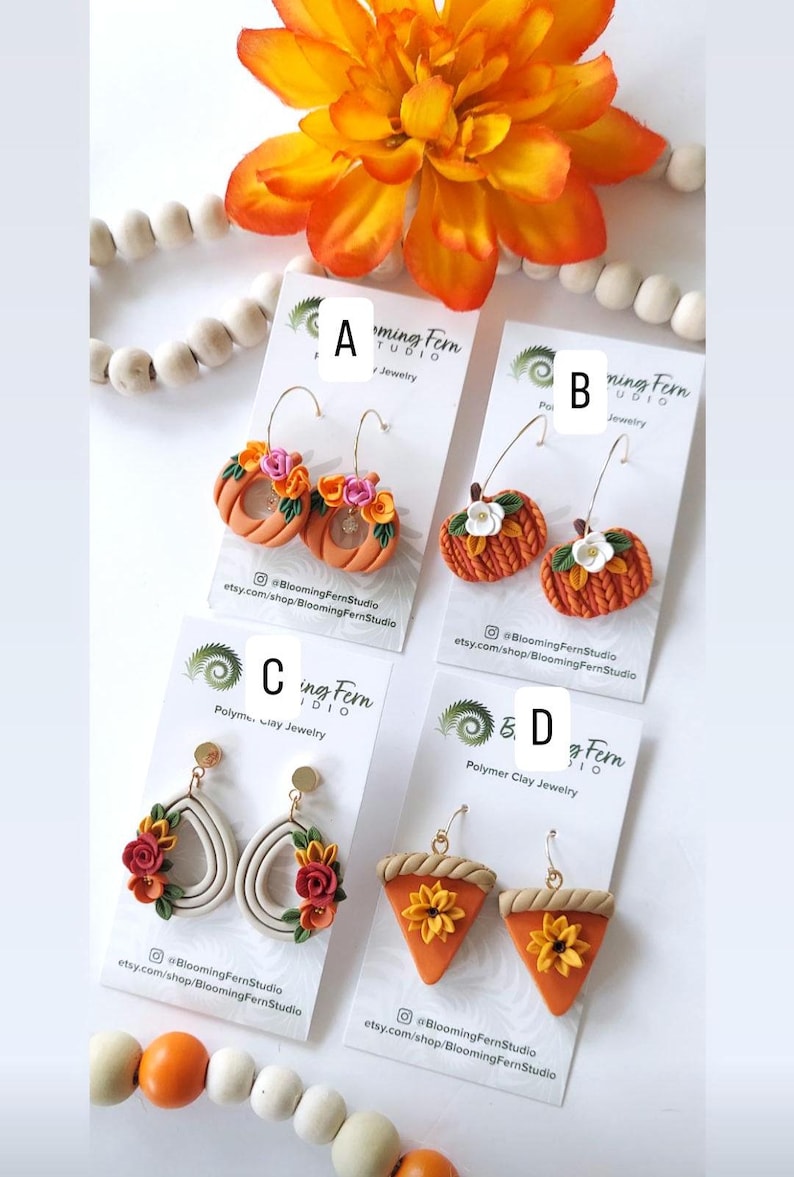 May include: A selection of polymer clay earrings in various designs. The earrings include orange pumpkin hoops, pumpkin-shaped earrings with white flowers, teardrop earrings with floral accents, and pie-shaped earrings with sunflowers. The earrings are displayed on white cards.