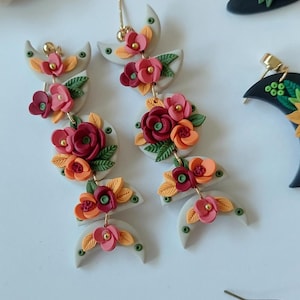 May include: Dangle earrings featuring stacked light gray crescent and circular clay shapes adorned with miniature red, orange, and pink flowers and green leaves. The earrings include gold-toned metal posts and connectors, showcasing a floral design.