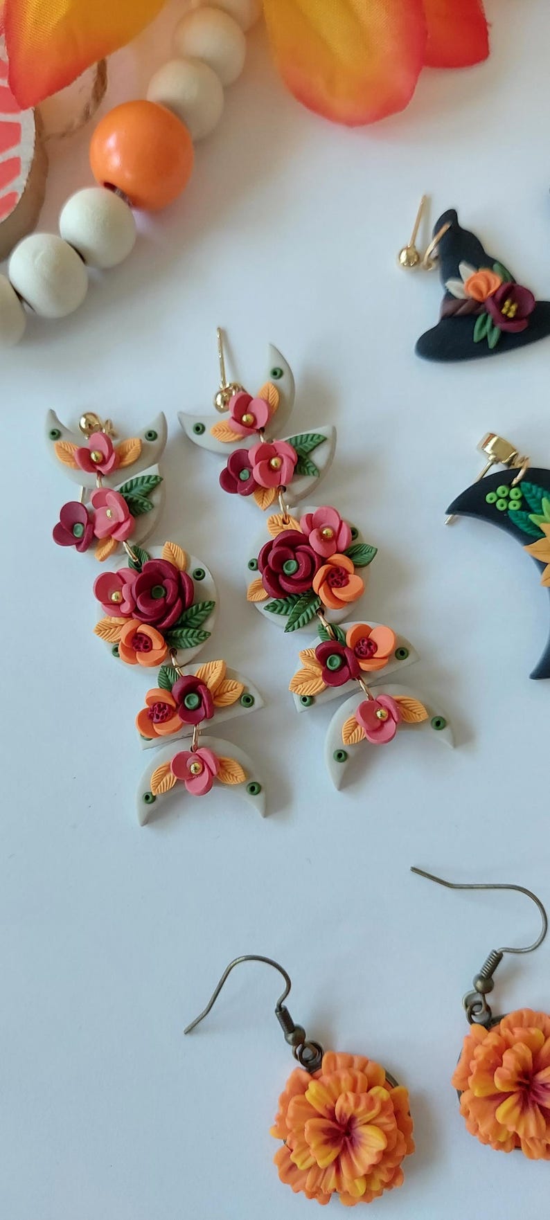 May include: Dangle earrings featuring stacked light gray crescent and circular clay shapes adorned with miniature red, orange, and pink flowers and green leaves. The earrings include gold-toned metal posts and connectors, showcasing a floral design.