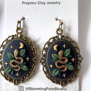 May include: A pair of oval-shaped earrings featuring a dark blue background with a gold snake, green leaves, a crescent moon, and small stars. The design is encased in an ornate, antique-style bronze metal frame with a hook-style earring wire.