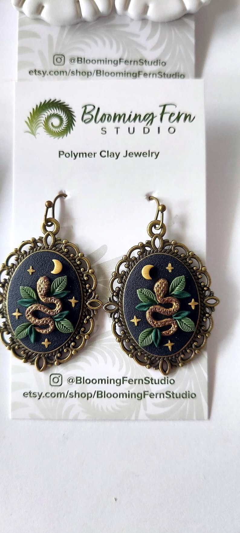 May include: A pair of oval-shaped earrings featuring a dark blue background with a gold snake, green leaves, a crescent moon, and small stars. The design is encased in an ornate, antique-style bronze metal frame with a hook-style earring wire.