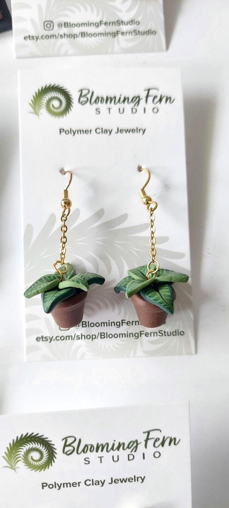 May include: A pair of dangle earrings featuring miniature potted plants made of polymer clay. Each earring has a brown clay pot with green leaves, suspended from a gold-toned metal chain and hook. The display card reads Blooming Fern Studio.