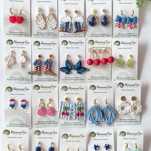 May include: A collection of twenty pairs of polymer clay earrings displayed on individual white branded cards. The designs include raspberries, oysters, books, anchors, bows, shells, cowboy boots, butterflies, cherries, grapes, popsicles, and stars.