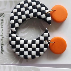 May include: A pair of earrings featuring a black and white checkered crescent shape attached to a solid orange circular stud. The jewelry is displayed on a white card labeled Blooming Fern Studio Polymer Clay Jewelry with social media handles.