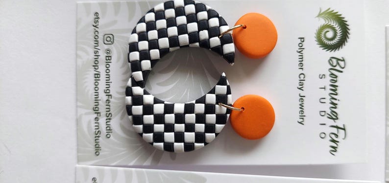 May include: A pair of earrings featuring a black and white checkered crescent shape attached to a solid orange circular stud. The jewelry is displayed on a white card labeled Blooming Fern Studio Polymer Clay Jewelry with social media handles.