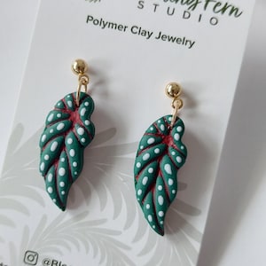 May include: A pair of dangle earrings featuring green polymer clay leaves with white polka dots and red veins, attached to gold-tone ball studs. The earrings are displayed on a white card labeled Blooming Fern Studio Polymer Clay Jewelry.
