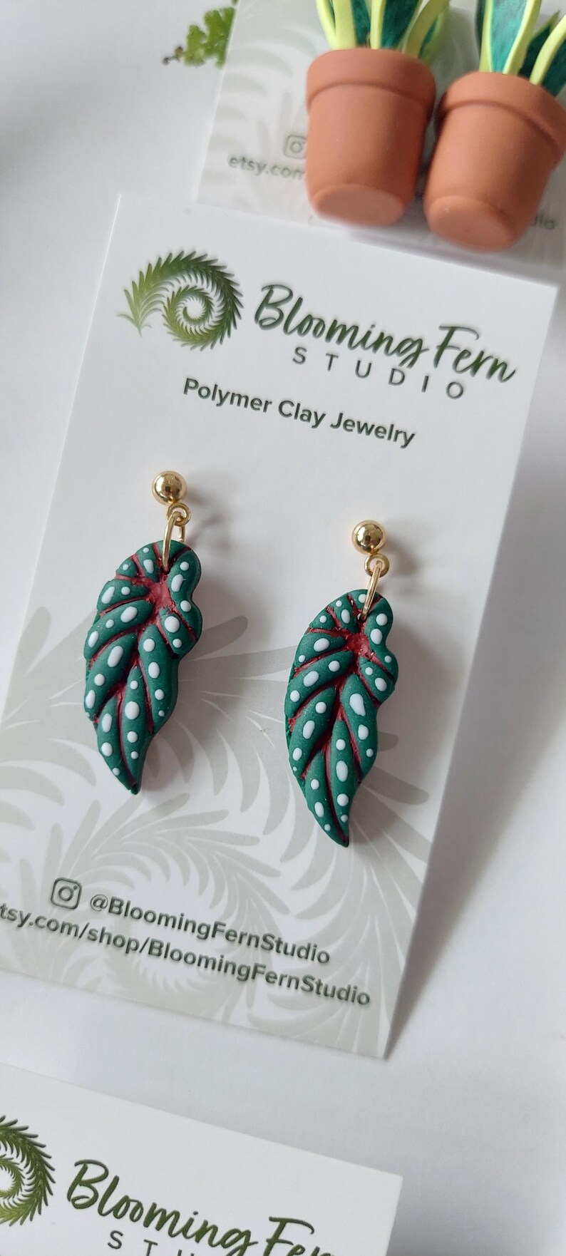 May include: A pair of dangle earrings featuring green polymer clay leaves with white polka dots and red veins, attached to gold-tone ball studs. The earrings are displayed on a white card labeled Blooming Fern Studio Polymer Clay Jewelry.