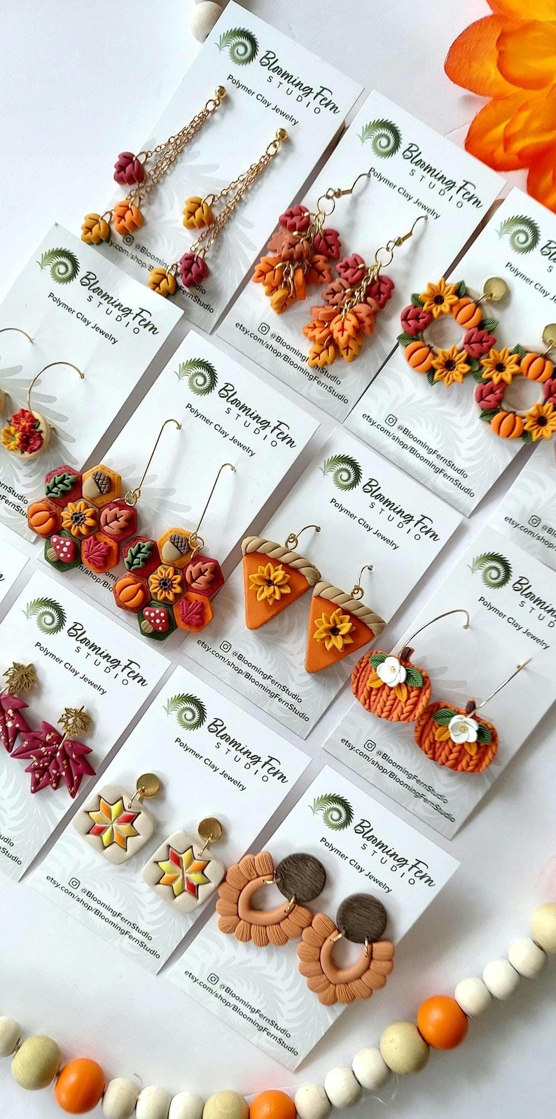 May include: A collection of polymer clay earrings in various fall-themed designs. The earrings feature leaf, pumpkin, and floral motifs in shades of orange, red, and yellow. Each pair is displayed on a white card with the Blooming Fern Studio logo.