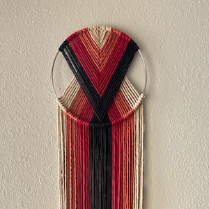 May include: A macrame wall hanging with a circular wooden frame and long strands of yarn in shades of red, orange, black, and cream.
