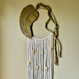 May include: A white and beige macrame wall hanging with a large green leaf shape at the top. The hanging has wooden beads and a gold hoop.