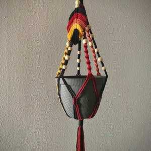 May include: A macrame plant hanger with a gray pot. The hanger is made with black, red, and yellow cord and has wooden beads.