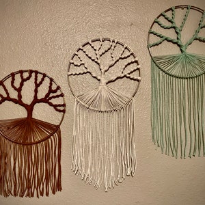 May include: Three macrame wall hangings in the shape of trees. The trees are made with brown, white, and green yarn. The hangings are all on a beige wall.