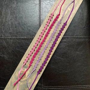 May include: Four friendship bracelets made with pink, purple, and lavender embroidery floss and small gold beads.