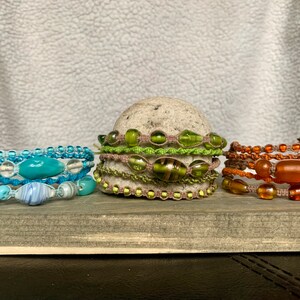 May include: Three bracelets made with colorful glass beads and cord. The bracelets are stacked on a wooden surface. The bracelet on the left has blue and white beads, the middle bracelet has green beads, and the bracelet on the right has orange and brown beads.